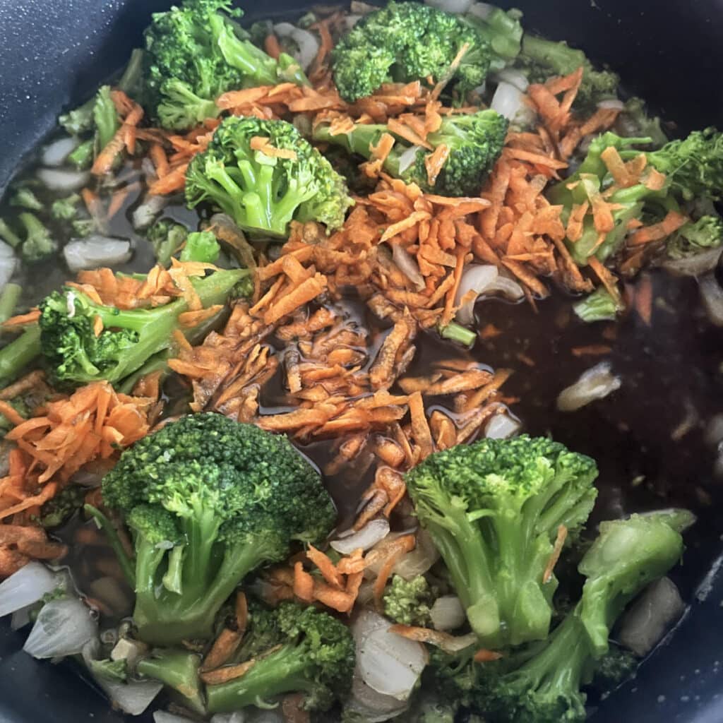 Broccoli florets and shredded carrots cooking in a sauce on the stove top.